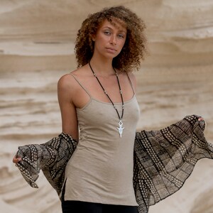 Op de afbeelding: Een vrouw met krullend bruin haar draagt een beige tanktop met een zilveren hangerketting en een zwart-wit gepatroneerde sjaal. Ze staat voor een zandige achtergrond.