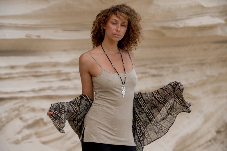 Op de afbeelding: Een vrouw met krullend bruin haar draagt een beige tanktop met een zilveren hangerketting en een zwart-wit gepatroneerde sjaal. Ze staat voor een zandige achtergrond.