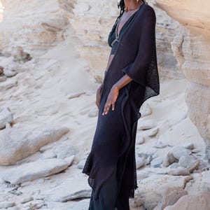 May include: A woman wearing a black knit shawl and matching pants. The shawl has a flowing design. The woman is also wearing a black top and sandals. The background is a light-colored rock formation.