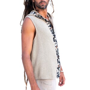 Op de afbeelding: Een mouwloos vest met capuchon in een lichtbeige kleur met een zwart-wit geometrisch patroon aan de zijkant. Het vest is gemaakt van een zachte, textuurrijke stof.
