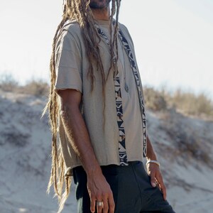 Op de afbeelding: Een man met lange dreadlocks draagt een beige shirt met korte mouwen met een zwart-wit geometrisch patroon. Hij draagt ook een zwarte broek en een zilveren ring aan zijn vinger.