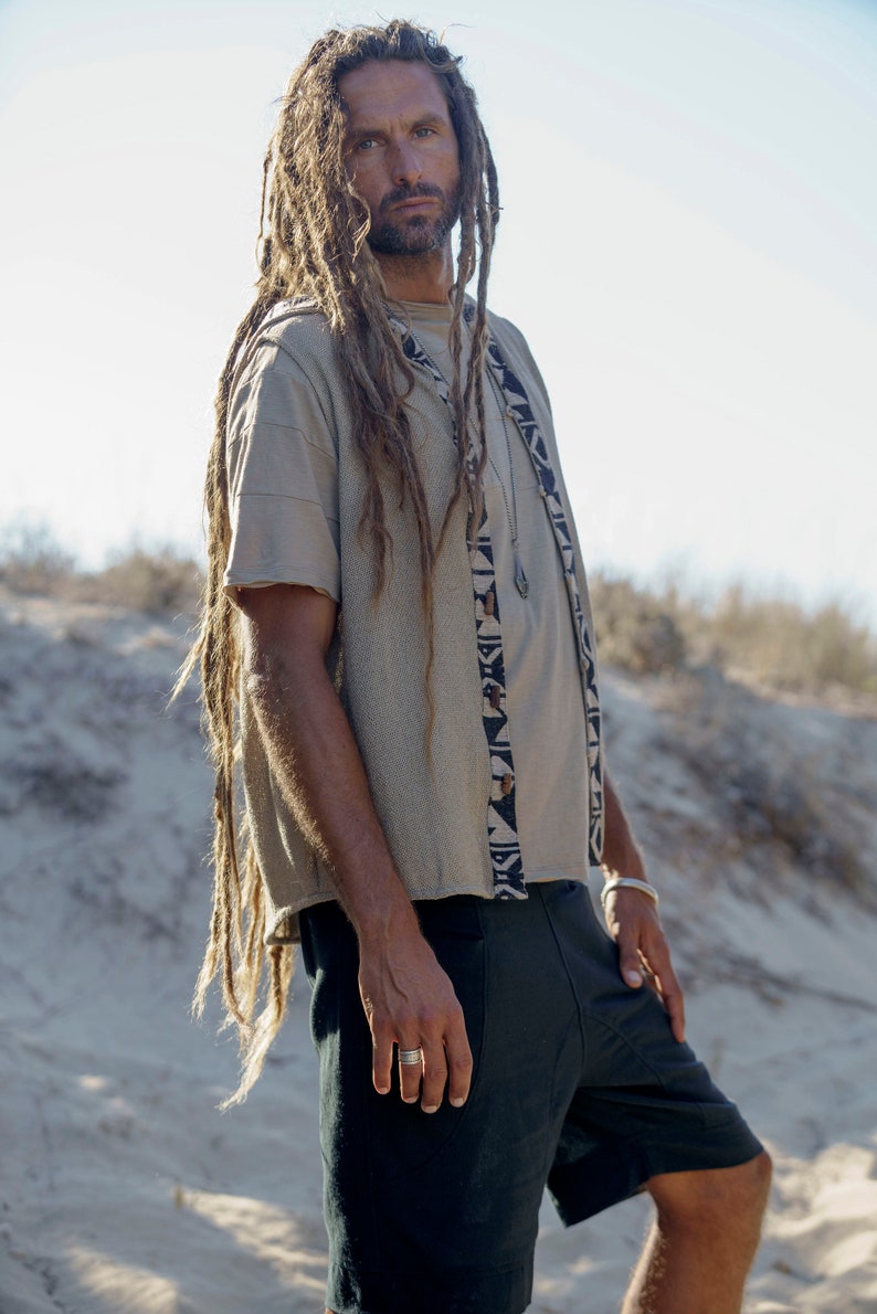 Op de afbeelding: Een man met lange dreadlocks draagt een beige shirt met korte mouwen met een zwart-wit geometrisch patroon. Hij draagt ook een zwarte broek en een zilveren ring aan zijn vinger.