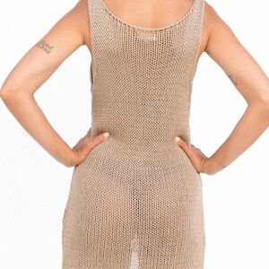Op de afbeelding: Een beige, mouwloze gebreide jurk. De jurk heeft een ronde hals en een licht uitlopende zoom. De gebreide stof heeft een getextureerde uitstraling. Het model staat met de handen in de zij.