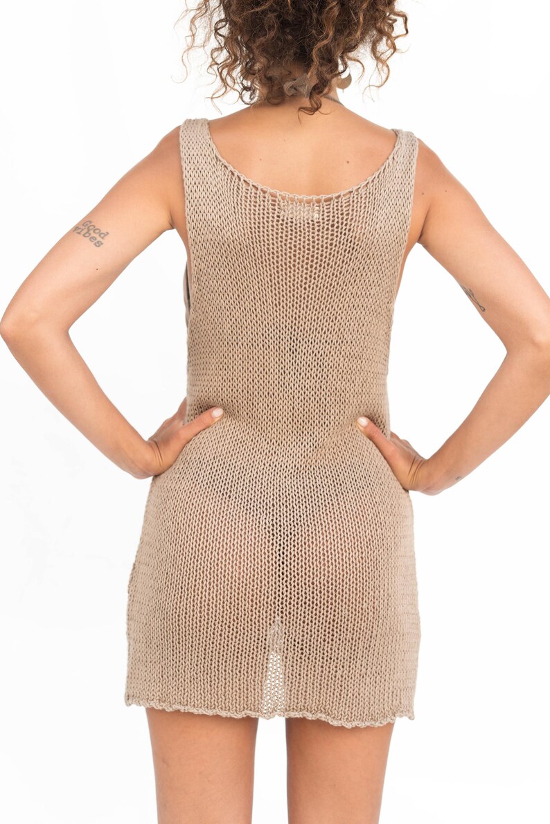 Op de afbeelding: Een beige, mouwloze gebreide jurk. De jurk heeft een ronde hals en een licht uitlopende zoom. De gebreide stof heeft een getextureerde uitstraling. Het model staat met de handen in de zij.