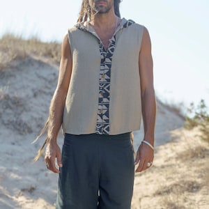 Puede incluir: Un hombre con rastas largas lleva una camisa sin mangas con capucha con un patr&oacute;n geom&eacute;trico y pantalones cortos azul oscuro. Est&aacute; de pie en una zona arenosa con hierba seca.