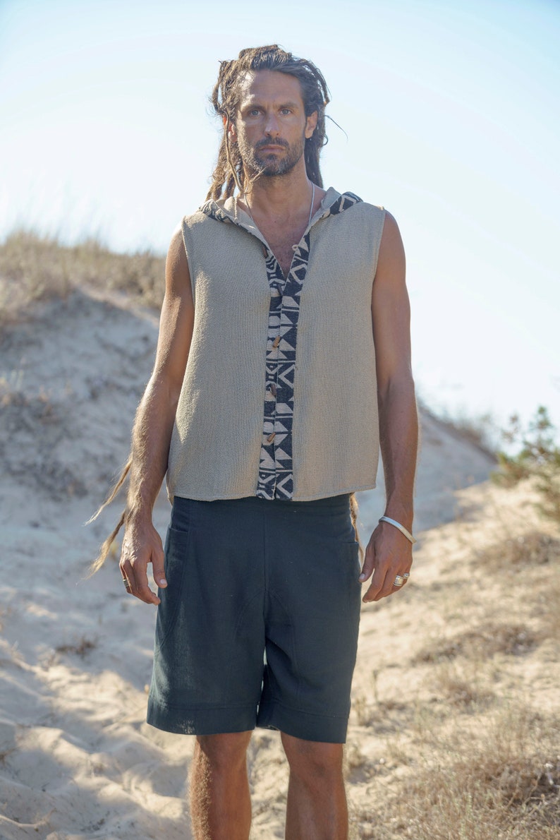 Puede incluir: Un hombre con rastas largas lleva una camisa sin mangas con capucha con un patr&oacute;n geom&eacute;trico y pantalones cortos azul oscuro. Est&aacute; de pie en una zona arenosa con hierba seca.