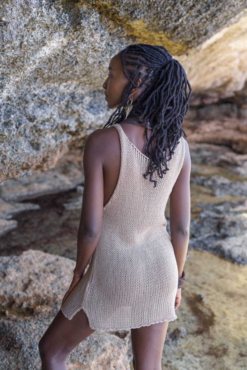 Op de afbeelding: Een beige, mouwloze gebreide jurk met een V-hals en zijsplitten. De jurk wordt gedragen door een persoon met donker haar en staat tegen een rotsachtige achtergrond. De jurk lijkt gemaakt van een zacht, ademend materiaal.