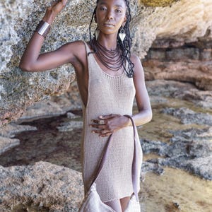 Op de afbeelding: Een vrouw draagt een beige gebreide jurk en een bijpassende schoudertas. Ze is versierd met gelaagde bruine kettingen, zilveren oorbellen en een zilveren armband. De achtergrond is een rotsachtige omgeving.