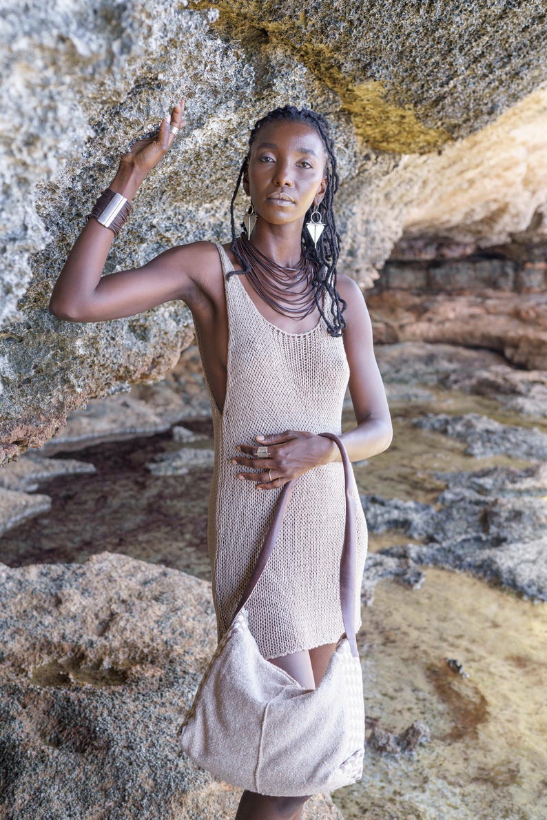 Op de afbeelding: Een vrouw draagt een beige gebreide jurk en een bijpassende schoudertas. Ze is versierd met gelaagde bruine kettingen, zilveren oorbellen en een zilveren armband. De achtergrond is een rotsachtige omgeving.