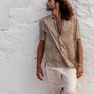 May include: A short-sleeved button-down shirt with a geometric pattern in shades of brown and tan. Paired with cream-colored shorts. The outfit is worn by a person leaning against a white textured wall.