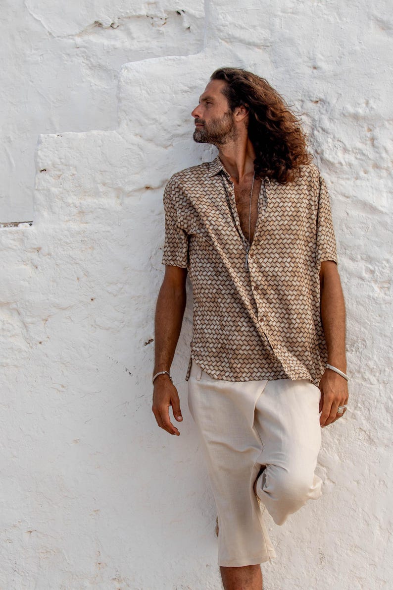 May include: A short-sleeved button-down shirt with a geometric pattern in shades of brown and tan. Paired with cream-colored shorts. The outfit is worn by a person leaning against a white textured wall.