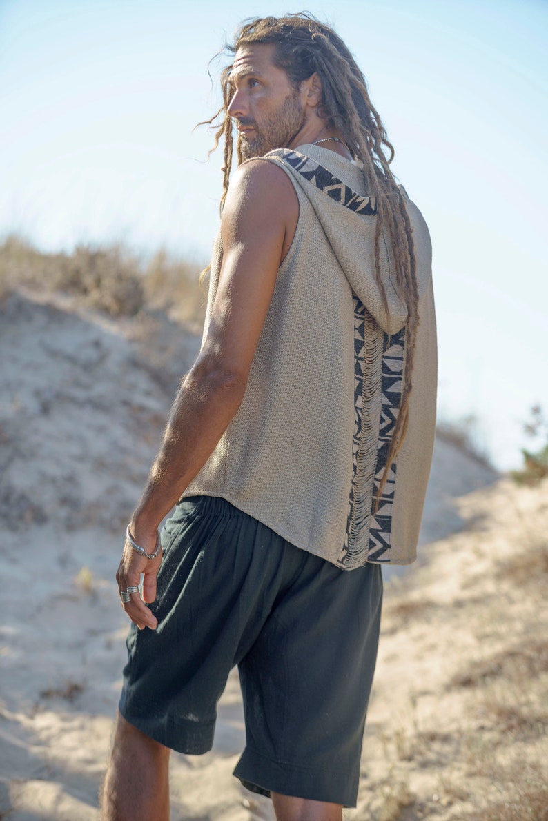 Op de afbeelding: Een man draagt een mouwloos hooded top met een geometrisch patroon en een zwarte broek. De top is beige met zwarte en witte geometrische ontwerpen. De man heeft lange dreadlocks.