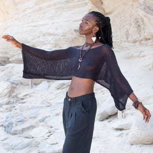 May include: A black, open-weave crop top with wide sleeves. The top is worn with dark trousers. The model is wearing jewellery, including a necklace and earrings. The background is a light-coloured, textured surface.