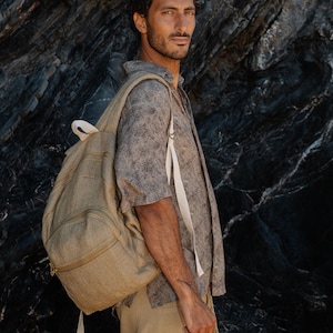 Può includere: Uno zaino intrecciato color beige con tasca frontale con cerniera e spallacci bianchi, indossato sopra una camicia grigia a maniche corte con motivo e pantaloni beige. Lo zaino presenta una struttura in tessuto testurizzato e una maniglia.