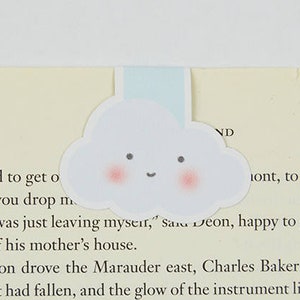 Pode incluir: Um marcador de página branco em forma de nuvem com um rosto sorridente e bochechas cor-de-rosa. O marcador de página está em uma página de um livro com texto que diz "I need to get o Can you drop m "I was just leaving mysert," sare Deon, happy to get t out of his mother's house. Deon drove the Marauder east, Charles Baker bes Night had fallen, and the glow of the instrument lights their faces. Baker looked at Deon, filling up his spac the wheel though the seat had been pushed far back. "You got some size on you," said Baker. "What you"