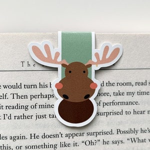 May include: A charming moose bookmark featuring a brown body, antlers, and a green tab. The bookmark is positioned on a book page, bringing a touch of nature-inspired charm to reading. The moose has a white outline.