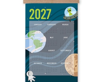 May include: A 2027 wall calendar featuring a space theme with a dark blue background, a green alien in a flying saucer, a depiction of Earth, a moon, and an astronaut. The calendar is held by wooden rails at the top and bottom with a hanging cord.