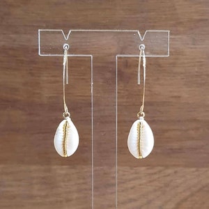May include: A pair of gold hoop earrings with white cowrie shell charms. The earrings are hanging from a clear acrylic display stand.