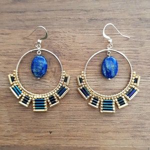 May include: A pair of hoop earrings with a silver finish. Each earring features a large, oval, deep blue gemstone suspended from a silver hook. The lower portion is adorned with gold, black, and teal beads, creating a decorative, semi-circular design.