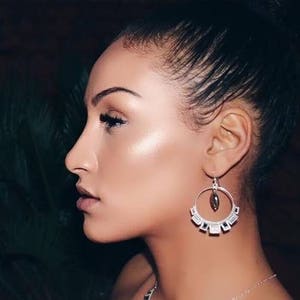 May include: A woman with dark hair wearing silver hoop earrings with a dark stone pendant. The earrings have a geometric design.