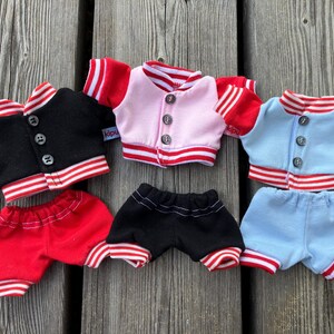 May include: Three sets of doll clothing are displayed. Each set includes a jacket and shorts. The jackets are black, pink, and light blue, with red and white striped trim. The shorts are red, black, and light blue, with matching trim.