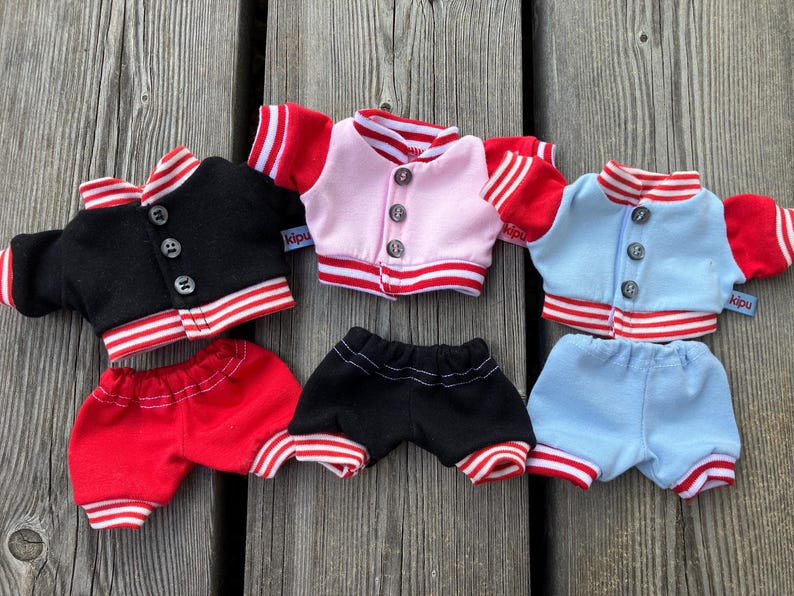May include: Three sets of doll clothing are displayed. Each set includes a jacket and shorts. The jackets are black, pink, and light blue, with red and white striped trim. The shorts are red, black, and light blue, with matching trim.