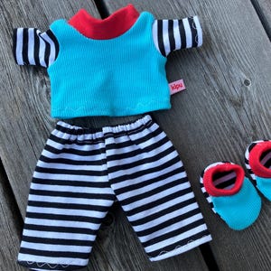 May include: A blue and white striped shirt with a red collar and a blue and white striped pair of pants. The shirt has a pink label that says "kipu". The outfit is accompanied by two blue and red striped shoes.