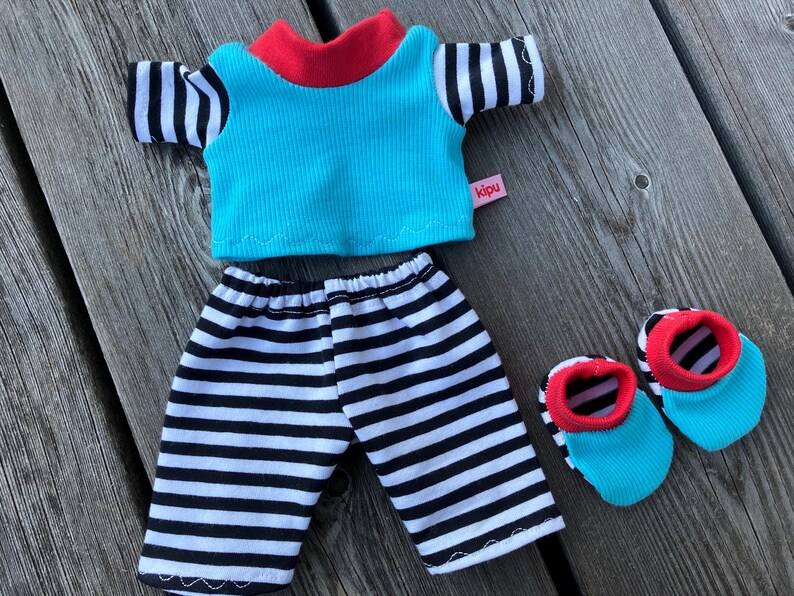 May include: A blue and white striped shirt with a red collar and a blue and white striped pair of pants. The shirt has a pink label that says "kipu". The outfit is accompanied by two blue and red striped shoes.