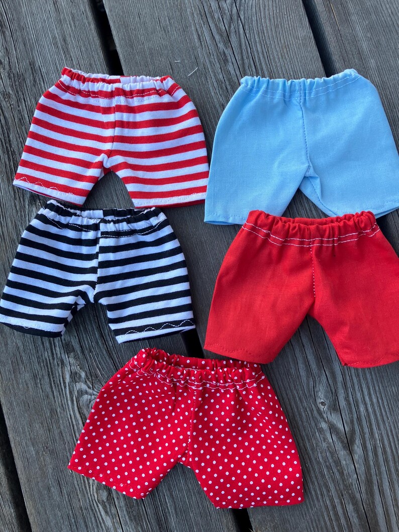 May include: Five pairs of shorts in different patterns. The shorts are red with white stripes, black with white stripes, light blue, red with white polka dots, and red with white stripes.