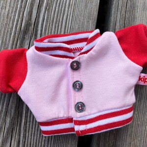 May include: A small pink and red varsity jacket with three buttons. The jacket has red sleeves with white stripes and a red and white striped collar and hem. A small pink tag with the word "kipu" is attached.