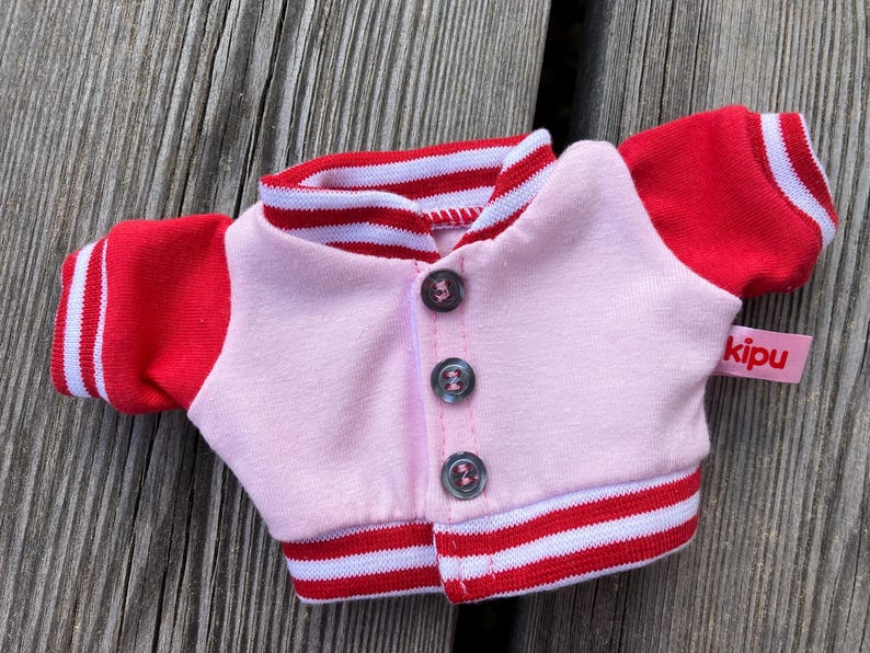 May include: A small pink and red varsity jacket with three buttons. The jacket has red sleeves with white stripes and a red and white striped collar and hem. A small pink tag with the word "kipu" is attached.