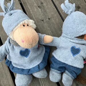 May include: Two plush toys wearing blue outfits with blue hearts on the front. The larger toy has a brown face and a blue hat with a knot on top. The smaller toy has a brown face and a blue hat with a fluffy brim.