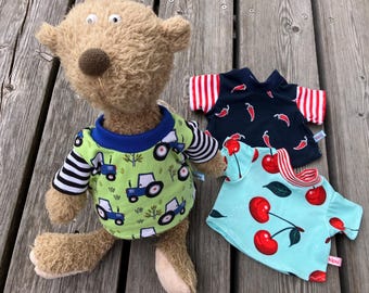 May include: A plush teddy bear wearing a green shirt with blue tractor graphics and black and white striped sleeves. Two additional shirts with fruit and chili pepper patterns are displayed nearby. The shirts are designed for a small stuffed animal.
