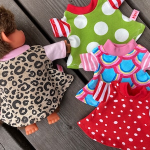 May include: A brown plush monkey doll wearing a leopard print dress with light pink sleeves, alongside three separate doll dresses: one green with white polka dots, one with a blue rainbow scale pattern, and one red with white polka dots, all featuring the label kipu.