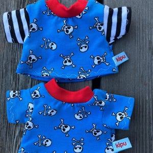 May include: Two blue t-shirts with a skull and crossbones pattern. The shirts have red trim and black and white striped sleeves. The shirts have a "kipu" label.