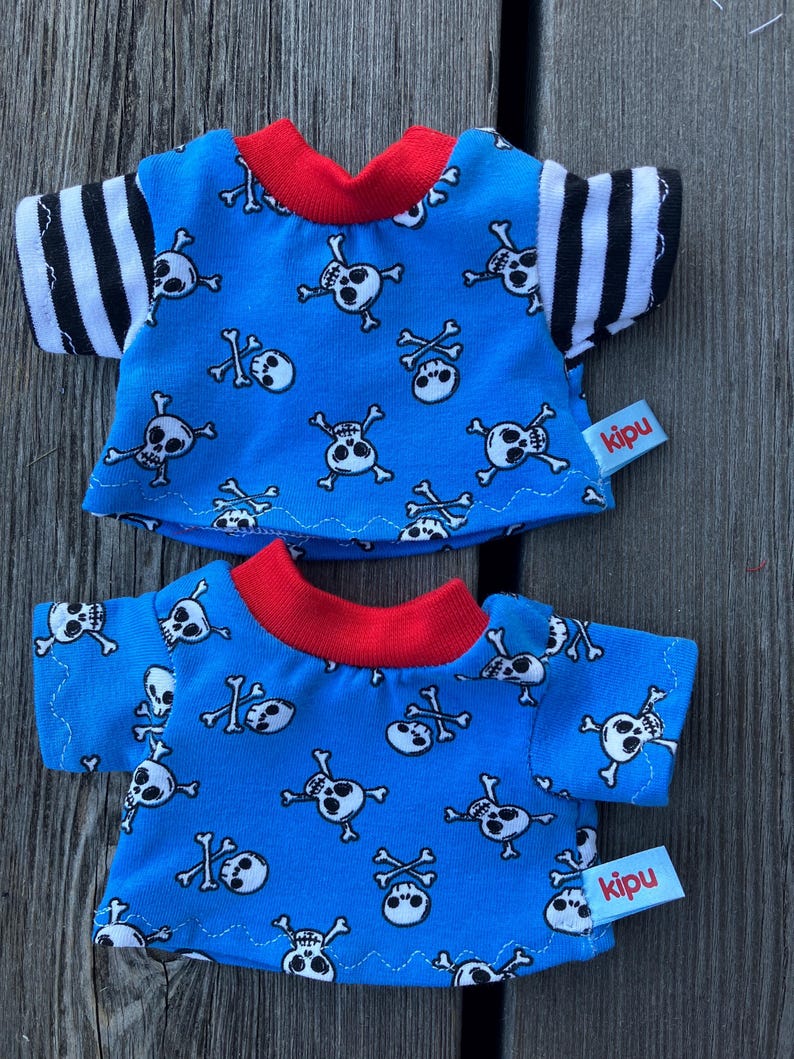 May include: Two blue t-shirts with a skull and crossbones pattern. The shirts have red trim and black and white striped sleeves. The shirts have a "kipu" label.