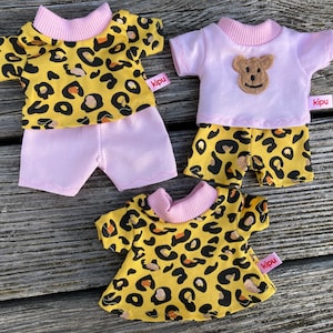 Könnte beinhalten: Drei Sätze Puppenkleidung mit einem gelben und schwarzen Leopardenmuster. Ein Satz enthält ein rosa Shirt und Shorts, ein anderer Satz enthält ein rosa Shirt und Shorts mit Leopardenmuster, und der dritte Satz ist ein Kleid. Alle Sätze haben einen rosa Besatz und ein "kipu"-Etikett.