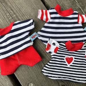Könnte beinhalten: Drei Miniatur-Puppenoutfits mit nautischem Thema. Die Outfits sind schwarz-weiß gestreift mit roten Akzenten. Ein Outfit hat ein rotes Herz auf der Vorderseite, ein anderes einen roten Kragen und das dritte eine rote Shorts. Jedes Outfit hat ein kleines blaues Etikett mit dem Wort "kipu" darauf.