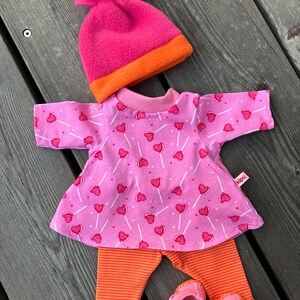 May include: A doll clothing set featuring a pink top with heart-shaped lollipop print, orange striped leggings, pink booties, and a pink and orange beanie. The set is designed for a doll and is made of soft fabric.