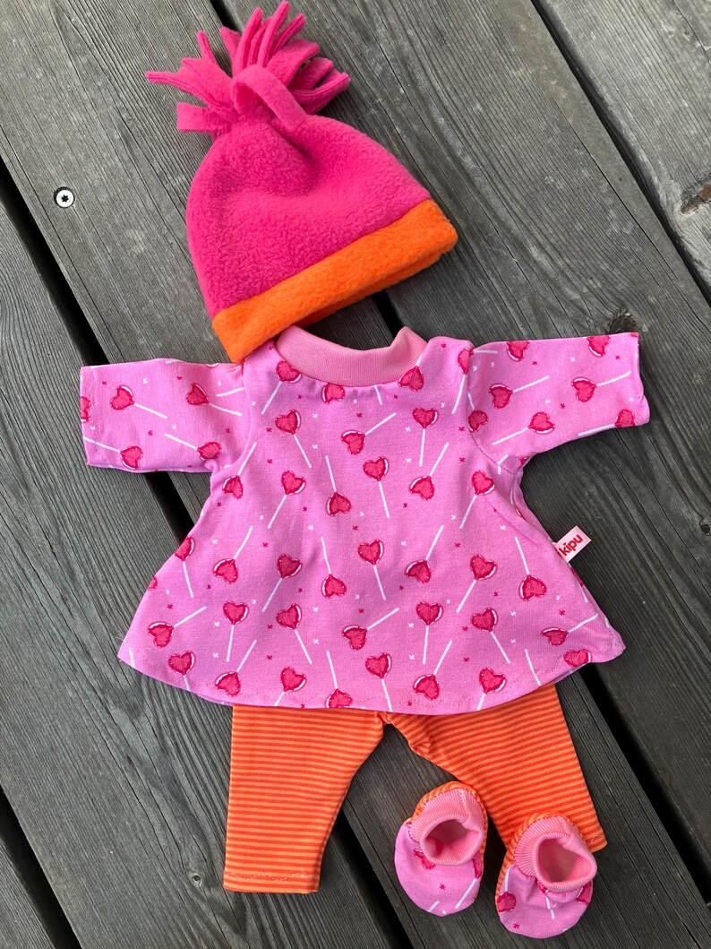 May include: A doll clothing set featuring a pink top with heart-shaped lollipop print, orange striped leggings, pink booties, and a pink and orange beanie. The set is designed for a doll and is made of soft fabric.