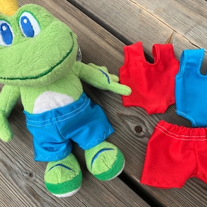 May include: A green frog soft toy wearing blue shorts, with red and blue swimsuits and red shorts. The frog has large blue eyes and a yellow crown. The toys are on a wooden surface.
