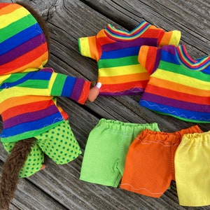 May include: A rainbow striped hooded sweatshirt, green polka dot pants, two rainbow striped t-shirts, and three pairs of shorts in green, orange, and yellow.