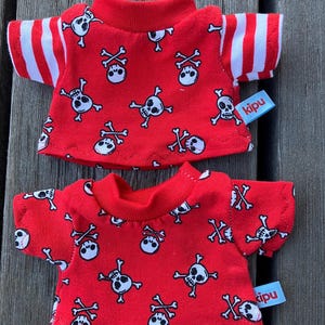 May include: Two red t-shirts with a white skull and crossbones pattern. The shirts have white stripes on the sleeves and a "kipu" label.