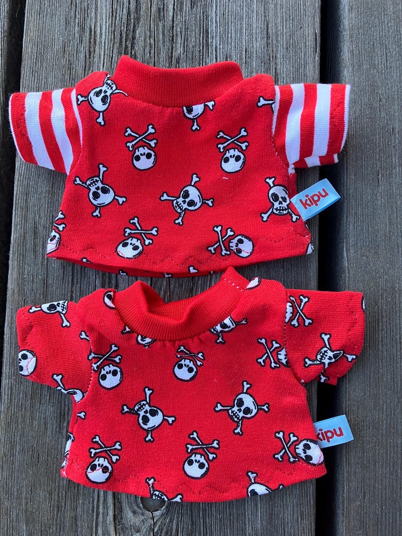 May include: Two red t-shirts with a white skull and crossbones pattern. The shirts have white stripes on the sleeves and a "kipu" label.
