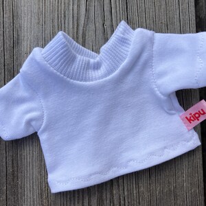 May include: A small, white t-shirt with short sleeves and a ribbed collar. A pink tag with the word "kipu" is sewn on the lower right side. The shirt is designed for a doll or small toy.
