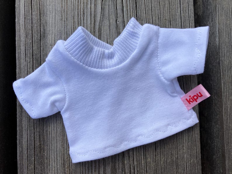 May include: A small, white t-shirt with short sleeves and a ribbed collar. A pink tag with the word "kipu" is sewn on the lower right side. The shirt is designed for a doll or small toy.
