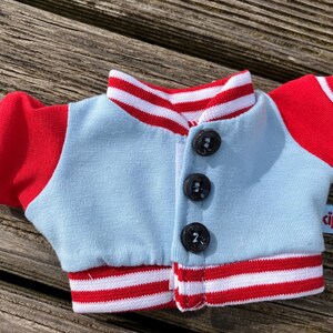 May include: A miniature light blue and red jacket with three black buttons. The jacket has red sleeves and a red and white striped collar and cuffs. A small blue tag with the word "cipu" is attached to the jacket.