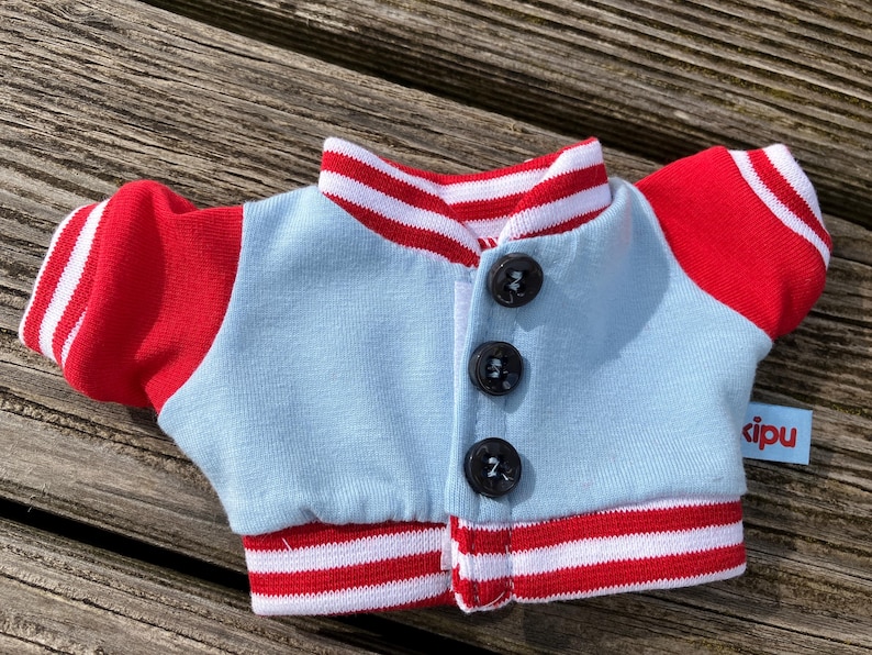 May include: A miniature light blue and red jacket with three black buttons. The jacket has red sleeves and a red and white striped collar and cuffs. A small blue tag with the word "cipu" is attached to the jacket.
