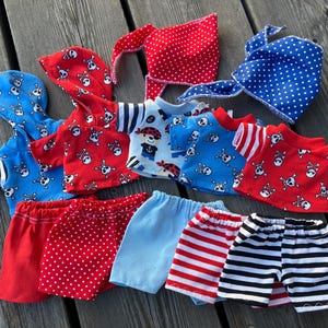 May include: A set of seven handmade doll clothes featuring a pirate theme. The set includes a red and white polka dot skirt, a blue and white striped skirt, a red and white striped skirt, a blue and white striped shirt, a red and white striped shirt, a red shirt with a skull and crossbones pattern, and a blue shirt with a skull and crossbones pattern.