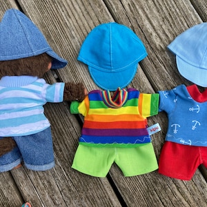 Könnte beinhalten: Drei Stofftiere, die verschiedene Outfits und Hüte tragen. Das erste Spielzeug ist ein brauner Affe, der einen blauen Jeanshut, ein blau-weiß gestreiftes Hemd und blaue Jeanshosen trägt. Das zweite Spielzeug trägt ein regenbogenfarbenes gestreiftes Hemd, grüne Shorts und einen blauen Hut. Das dritte Spielzeug trägt ein blaues Hemd mit weißen Ankern, rote Shorts und einen hellblauen Hut.